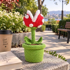 Puede incluir: Una planta de ganchillo en una maceta verde con una cabeza roja de puntos blancos y dientes blancos, un tallo verde y dos hojas. La planta está junto a un vaso de café beige con el texto Good Days Ahead y un libro con Small Steps Big Changes.