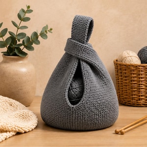 May include: A grey knitted yarn bowl featuring a unique Japanese knot bag design with a single handle loop. The structure is crafted from a textured knit fabric, creating a rounded pouch shape that securely holds a ball of yarn inside the center opening.