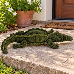 May include: A dark green crocheted alligator stuffed animal featuring a textured back with raised ridges, a long tail, four short legs, and a white underside along the jawline. The creature has small yellow eyes and a detailed, knotted yarn surface.