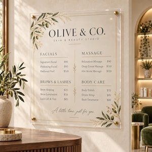 Op de afbeelding: Een rechthoekig bord van helder acryl voor Olive & Co. Skin & Beauty Studio, bevestigd met gouden schroeven. Het toont een lijst met diensten zoals gezichtsbehandelingen, massages, wenkbrauwen, wimpers en lichaamsverzorging, versierd met olijftakken.