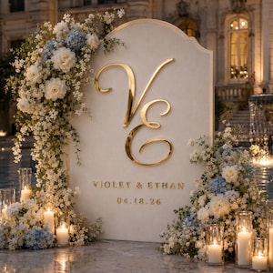 May include: An arched cream-coloured wedding sign featuring large gold script letters V and E, with the text VIOLET & ETHAN and the date 04.18.26. The sign is decorated with white and blue flowers and surrounded by glass pillar candles.