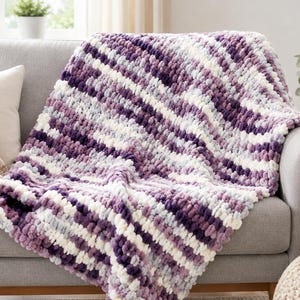 May include: A chunky knit throw blanket featuring a variegated pattern of deep purple, light lavender, and white yarn. The blanket is constructed with thick, soft loops of chenille-style yarn, creating a textured, plush surface with diagonal stripes.