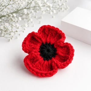 Può includere: Un fiore lavorato all'uncinetto a mano con petali rosso vivace disposti in un motivo circolare attorno a un centro nero testurizzato. La lavorazione del filato mostra punti visibili e un aspetto morbido e tridimensionale.