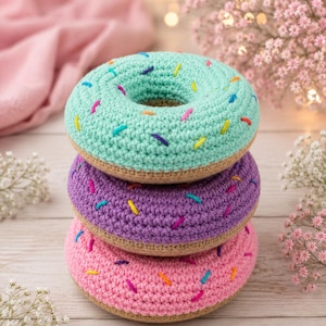 Pode incluir: Uma pilha de três brinquedos de crochê em forma de donut. O donut do topo é verde-menta, o do meio é roxo e o de baixo é rosa. Cada donut possui uma base bege e é decorado com pequenos pontos de fio coloridos que representam granulados amarelos, azuis e roxos.