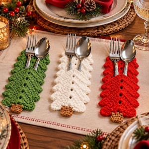 May include: Three crochet silverware holders shaped like Christmas trees in green, white, and red. Each holder features a small brown trunk at the base and contains a silver metal fork and spoon. The holders are arranged side by side on a table.