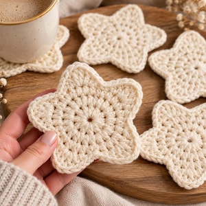 Puede incluir: Un juego de posavasos de ganchillo en forma de estrella de color blanco roto, hechos con hilo grueso. Cada posavasos presenta un centro circular con puntos radiantes que forman cinco puntas distintas con un diseño consistente.