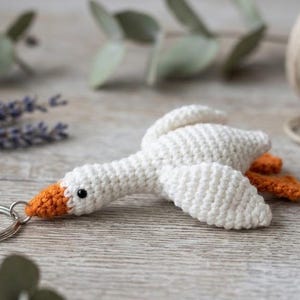 Könnte beinhalten: Ein weißer, gehäkelter Gänse-Schlüsselanhänger mit orangefarbenem Schnabel und orangefarbenen Füßen. Die Gans hat ein kleines schwarzes Perlenauge und ist an einem runden silbernen Metallring befestigt. Die Häkelarbeit zeigt deutliche Maschen am Körper.