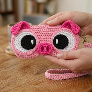 Peut inclure: Une pochette crochetée rose en forme de visage de cochon, avec de grands yeux blancs et noirs, un petit museau rose et deux petites oreilles roses. La pochette comprend une dragonne en crochet rose assortie fixée par un mousqueton en métal argenté.