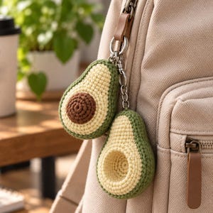 May include: A pair of crocheted avocado keychains attached to a beige canvas backpack. One avocado features a dark brown center pit, while the other shows a hollowed-out center. Both are crafted with light yellow and green yarn, connected by a silver metal ring.