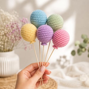 Op de afbeelding: Een set van vijf kleine, ronde, gehaakte garenballonnen in pasteltinten van geel, lichtblauw, lavendel, mintgroen en roze. Elke bol is bevestigd aan een dun, recht houten stokje, wat een tros van kleurrijke, getextureerde ballonvormen creëert.
