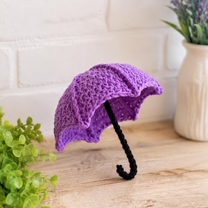 May include: A small, decorative crochet umbrella featuring a vibrant purple canopy with a textured, stitched pattern and a curved black handle. The umbrella is constructed from yarn, showcasing detailed needlework throughout the canopy and handle.