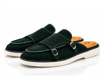 May include: A pair of dark green suede mules featuring a double monk strap design with silver-toned metal buckles. The footwear has a thick white rubber sole and a tan-colored interior footbed with a small black logo printed on the heel area.