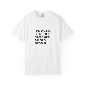 Puede incluir: Una camiseta blanca de manga corta con el texto negro IT'S WEIRD BEING THE SAME AGE AS OLD PEOPLE. centrado en el pecho. La prenda tiene un cuello redondo y un corte estándar, con una pequeña etiqueta de marca visible en el cuello.