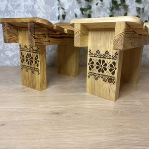 May include: Two light-coloured wooden stools with a curved seat design. Each stool features a dark brown laser-engraved floral pattern on the front leg, showcasing a repeating geometric motif. The wood grain is visible throughout the structure.