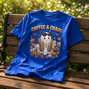 May include: A royal blue t-shirt featuring a graphic of a ghost wearing sunglasses and a leopard print scarf, holding a coffee cup. The design includes the text COFFEE & CHAOS, surrounded by autumn leaves, small pumpkins, and bats in a fall theme.