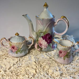 Puede incluir: Un juego de té de porcelana que consta de una tetera con tapa, un azucarero con tapa y una jarra de leche. Cada pieza presenta una base de color azul pálido y blanco con rosas pintadas a mano, hojas verdes y detalles en bordes dorados.