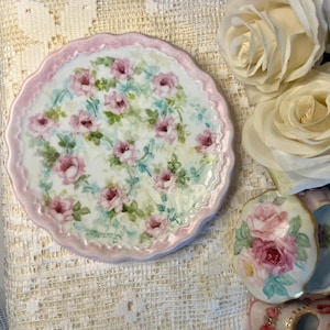 May include: A round porcelain plate with a scalloped edge, featuring a hand-painted pattern of pink roses and green leaves on a white background, with a soft pink border. A small matching lidded trinket box with floral motifs sits nearby.