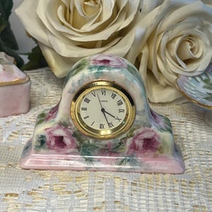May include: A ceramic mantel clock with a curved top and a rectangular base, featuring a hand-painted pink rose floral pattern on a cream background. The clock face is white with black Roman numerals and a gold-tone circular bezel.