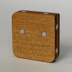 Könnte beinhalten: Ein quadratischer Holzblock mit abgerundeten Ecken und einer ausgeprägten natürlichen Holzmaserung. Der Block ist mit sechs kreisförmigen, silberfarbenen Metallscheiben versehen, zwei auf der Vorderseite und vier an den Seitenkanten, jeweils mit einem dünnen Kupferring.