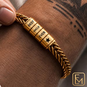 May include: A gold-toned wheat chain bracelet featuring three cylindrical beads. The beads are engraved with the text BEST, UNCLE, and EVER, followed by a black star symbol. The bracelet has a polished metallic finish and a woven link design.