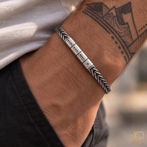 Peut inclure: Un bracelet argenté composé d'une chaîne tressée et de quatre perles cylindriques. Chaque perle est gravée de symboles en code Morse noirs. Le bracelet présente une finition métallique et est porté au poignet, révélant un motif de maillons détaillé.