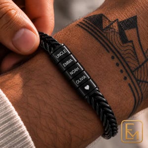 Könnte beinhalten: Ein schwarzes Weizenkettenarmband aus Metall mit fünf zylindrischen Perlen. Jede Perle ist mit weißem Text graviert, der UNCLE, EMMA, NOAH, OLIVIA und ein kleines Herzsymbol zeigt. Das Armband hat eine polierte Oberfläche und einen Verschluss.