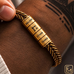 Könnte beinhalten: Ein goldfarbenes Metallkettenarmband mit einer zylindrischen Mittelperle, die mit dem Text BUILDER, einem Kransymbol, dem Namen ETHAN und dem Wort THANKS graviert ist. Das Armband hat ein geflochtenes Weizenkettenmuster und ein poliertes Finish.