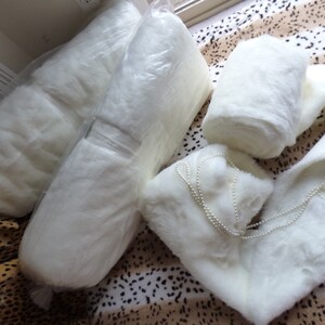 May include: Two white faux fur fabric rolls, one rolled up and one flat, with a white faux fur scarf and a string of white pearls.