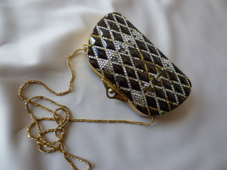 Peut inclure: Un sac de soir&eacute;e noir et or avec une bandouli&egrave;re cha&icirc;ne. Le sac est recouvert de strass et pr&eacute;sente un motif en diamant.