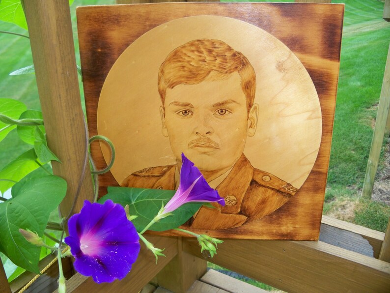 Peut inclure: Un portrait d'un homme en uniforme militaire grav&eacute; au pyrograve sur du bois, encadr&eacute; dans un cadre en bois. Une fleur de gloire du matin violette est au premier plan.