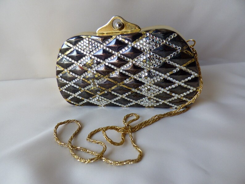 Peut inclure: Un sac de soir&eacute;e noir et argent avec une bandouli&egrave;re en cha&icirc;ne dor&eacute;e. Le sac est recouvert de strass et pr&eacute;sente un motif matelass&eacute;.