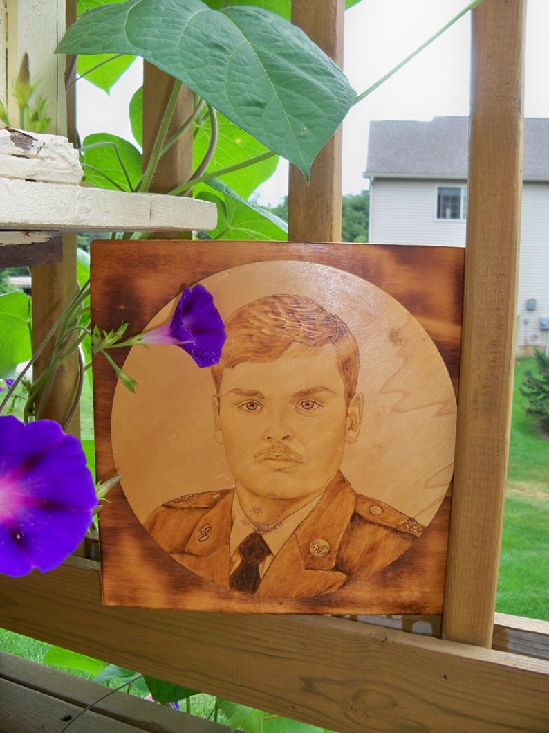 Peut inclure: Un portrait grav&eacute; au pyrograveur d'une personne en uniforme militaire sur un fond brun clair. Le portrait est encadr&eacute; dans un cadre en bois. Une fleur de gloire du matin violette est au premier plan.
