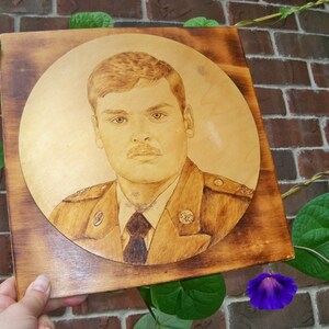 Peut inclure: Une plaque en bois avec une d&eacute;coupe circulaire repr&eacute;sentant le portrait d'un homme en uniforme militaire. L'homme porte une cravate fonc&eacute;e et a les cheveux courts bruns. La plaque a un grain de bois brun clair.