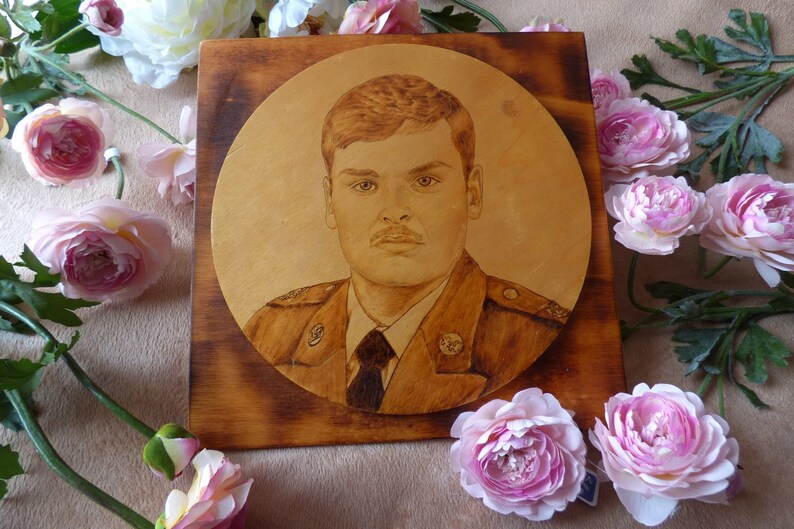 Peut inclure: Une plaque en bois avec une image br&ucirc;l&eacute;e circulaire d'un homme en uniforme militaire. La plaque est entour&eacute;e de fleurs roses.