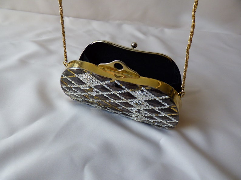 Peut inclure: Un sac &agrave; main de soir&eacute;e avec une bandouli&egrave;re en cha&icirc;ne dor&eacute;e, un int&eacute;rieur en velours noir et un ext&eacute;rieur transparent orn&eacute; de bijoux. Le sac est d&eacute;cor&eacute; d'un motif g&eacute;om&eacute;trique de strass blancs.