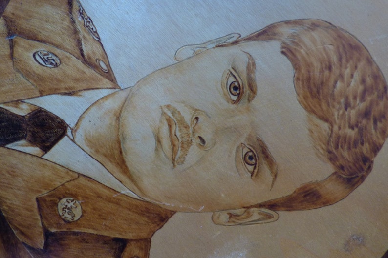 Peut inclure: Un portrait pyrograv&eacute; d'une personne en uniforme militaire. La personne a les cheveux bruns et regarde de c&ocirc;t&eacute;. Le portrait est r&eacute;alis&eacute; dans des tons de brun et de beige.