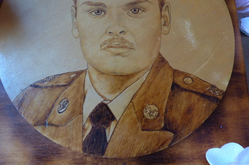 Peut inclure: Un portrait pyrograv&eacute; d'une personne en uniforme militaire. La personne porte un uniforme brun avec une cravate noire et un badge sur le c&ocirc;t&eacute; gauche de la poitrine. L'arri&egrave;re-plan est brun clair.