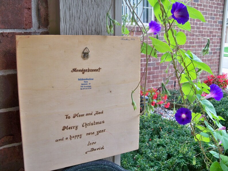 Peut inclure: Une pancarte en bois avec un message manuscrit qui dit "To Mom and Dad Merry Christmas and a happy new year love David". La pancarte est accroch&eacute;e &agrave; un mur de briques avec une vigne de fleurs de gloire du matin violettes qui pousse le long du mur.