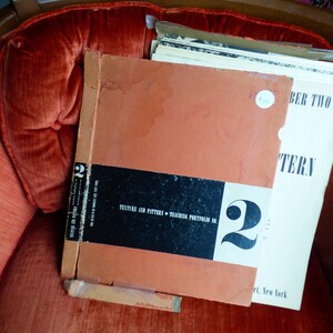 Op de afbeelding: Een vintage oranje en zwart boek getiteld "TEXTURE AND PATTERN - TEACHER'S PORTFOLIO 2" met het nummer "2" in groot zwart lettertype. Het boek ligt op een stapel wit papier met de tekst "NUMBER TWO" en "PATTERN" zichtbaar.
