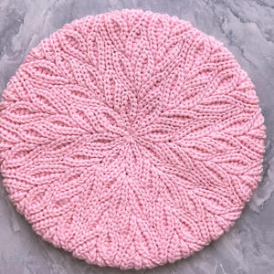May include: A circular, light pink knitted beret featuring a textured, radiating leaf-like pattern that originates from the center and extends outward to the edges. The knit construction is uniform, displaying a consistent stitch density throughout.