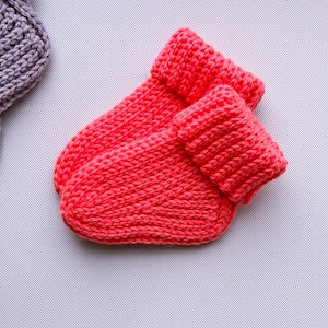 May include: A pair of bright coral-coloured knit booties featuring a thick, folded cuff and a textured, chunky yarn construction. The booties are crafted with a visible stockinette stitch pattern and have a rounded toe shape, showcasing a vibrant, solid colour throughout the entire design.