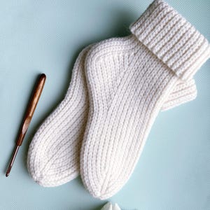 May include: A pair of off-white knitted socks featuring a ribbed cuff and a textured knit pattern, accompanied by a dark brown wooden crochet hook positioned to the left of the socks, showcasing the craft and construction of the footwear.