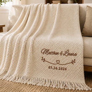 May include: A cream-coloured textured knit throw blanket featuring fringe trim along the bottom edge. The blanket is embroidered with the brown text "Matthew & Laura", a decorative heart and vine motif, and the date "07.26.2026" in a script font.