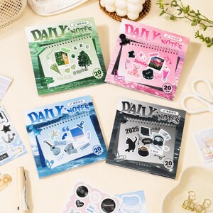 May include: Four square spiral-bound Daily Notes pads with 20 pages each. The green pad features nature stickers, the pink pad displays cute icons, the blue pad shows sea-themed graphics, and the black pad includes music and cat illustrations.