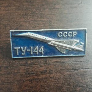 Pode incluir: Um broche retangular de metal com fundo de esmalte azul e um gráfico de avião supersônico em tom prateado. O texto CCCP está impresso no canto superior direito e o texto TY-144 está impresso no canto inferior esquerdo do broche.
