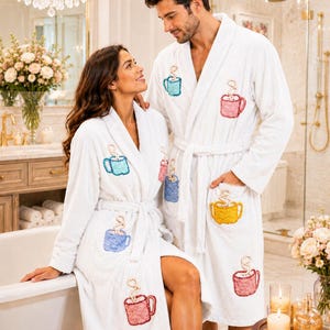 Puede incluir: Dos batas de baño blancas de felpa con diseños bordados de tazas coloridas con vapor saliendo de cada una. Las tazas aparecen en tonos azul, rosa y amarillo, distribuidas por el frente de las batas, las cuales incluyen cinturones a juego.