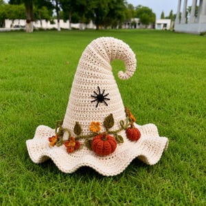Op de afbeelding: Een crèmekleurige gehaakte heksenhoed met een gebogen punt, voorzien van een zwarte spin-applicatie op de kroon en een decoratieve rankenslinger rond de basis, versierd met kleine oranje pompoenen, groene bladeren en gele en oranje bloemen.