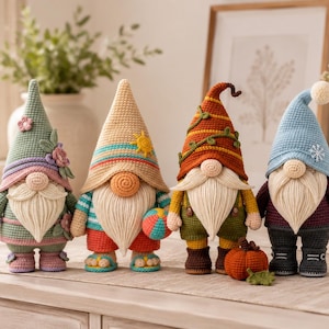 Peut inclure: Quatre figurines de gnomes au crochet représentant les différentes saisons. Le premier est vert avec des fleurs, le deuxième est beige avec un soleil et un ballon de plage, le troisième est orange avec des vignes et une citrouille, et le quatrième est bleu avec un flocon de neige.