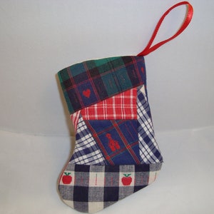 May include: A small, patchwork Christmas stocking with a red ribbon loop. The stocking is made of various plaid fabrics in red, blue, green, and white. It features a red heart, a small schoolhouse, and red apples.