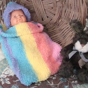 May include: A baby doll wrapped in a rainbow striped fuzzy blanket, lying in a wicker basket. A black plush bunny with a white face and a black and white checkered bow sits next to the doll.